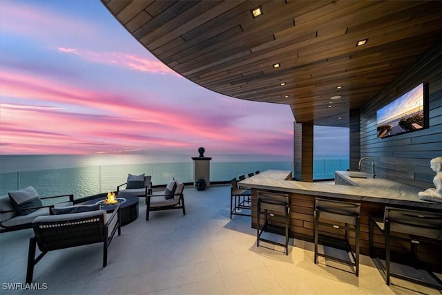 patio terrace at dusk with a water view, an outdoor fire pit, and an outdoor wet bar