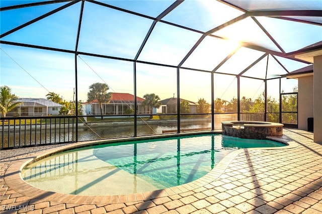 pool at dusk with an in ground hot tub, a water view, a patio area, and glass enclosure