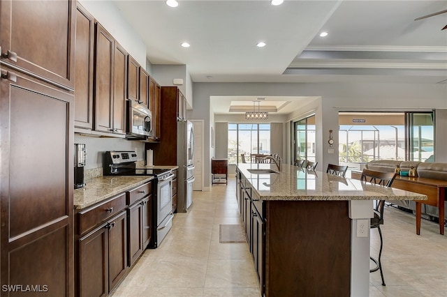 kitchen with a breakfast bar area, light stone counters, a tray ceiling, a center island with sink, and appliances with stainless steel finishes