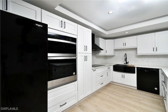 kitchen with wall chimney exhaust hood, a tray ceiling, sink, black appliances, and white cabinets