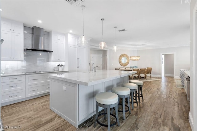 kitchen with white cabinetry, wall chimney exhaust hood, an island with sink, pendant lighting, and light hardwood / wood-style floors