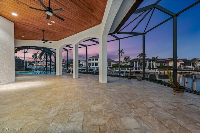 patio terrace at dusk featuring glass enclosure, ceiling fan, and a water view