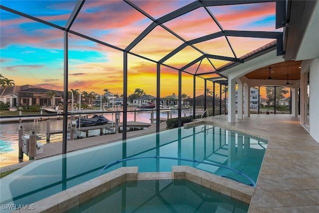 pool at dusk featuring glass enclosure, a water view, a boat dock, ceiling fan, and an in ground hot tub