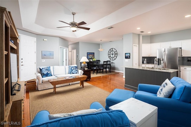 living room with a raised ceiling, ceiling fan, sink, and light wood-type flooring