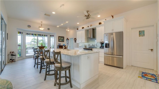 kitchen featuring white cabinets, decorative light fixtures, wall chimney range hood, stainless steel appliances, and a center island with sink