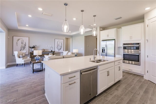 kitchen featuring a kitchen island with sink, a raised ceiling, appliances with stainless steel finishes, white cabinets, and sink