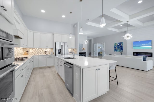 kitchen with coffered ceiling, a large fireplace, stainless steel appliances, white cabinets, and an island with sink