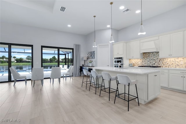 kitchen with white cabinetry, hanging light fixtures, premium range hood, a towering ceiling, and a center island with sink