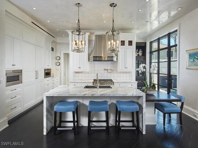 kitchen with a large island with sink, oven, sink, wall chimney exhaust hood, and white cabinetry