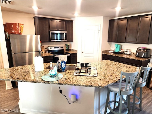 kitchen with dark wood-type flooring, an island with sink, appliances with stainless steel finishes, light stone counters, and dark brown cabinetry