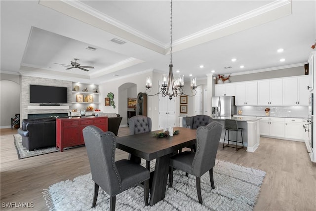 dining room featuring ceiling fan with notable chandelier, a tray ceiling, crown molding, light hardwood / wood-style flooring, and a fireplace