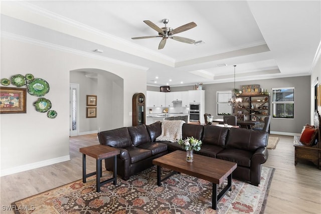 living room with ceiling fan with notable chandelier, light wood-type flooring, crown molding, and a tray ceiling