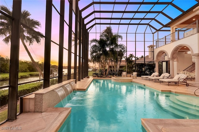 pool at dusk featuring pool water feature, glass enclosure, an in ground hot tub, and a patio