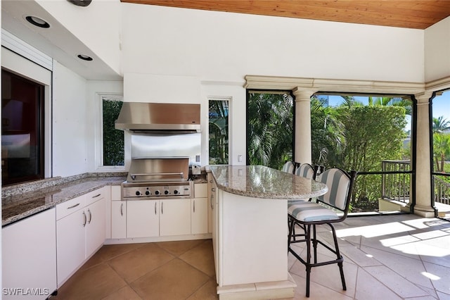 kitchen featuring light stone counters, ventilation hood, light tile patterned floors, white cabinetry, and a breakfast bar area