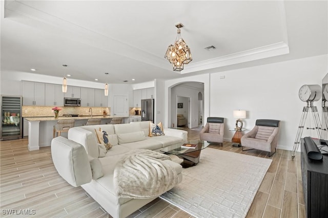 living room featuring sink, wine cooler, a notable chandelier, crown molding, and a tray ceiling