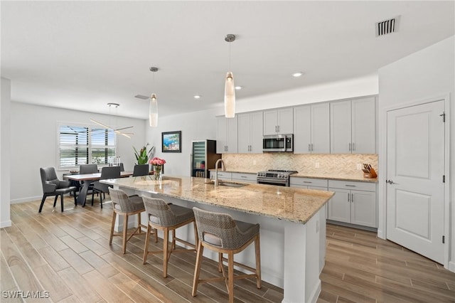 kitchen featuring light stone counters, stainless steel appliances, a kitchen island with sink, sink, and hanging light fixtures