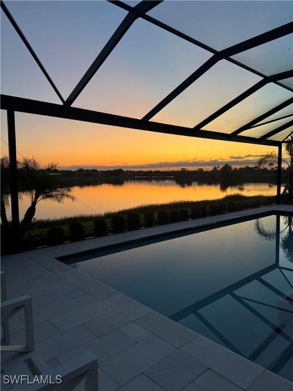 pool at dusk featuring a water view, a patio area, and a lanai