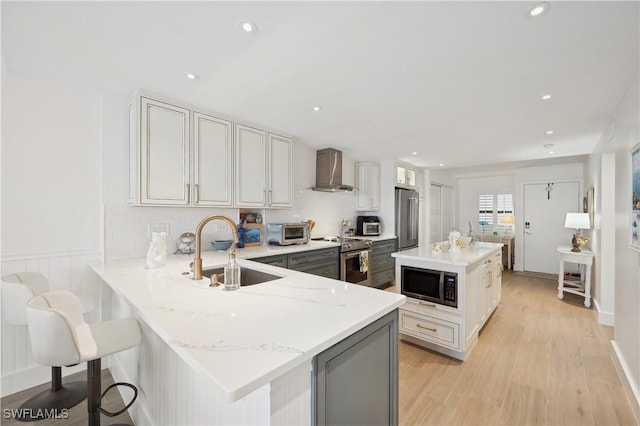 kitchen featuring wall chimney range hood, sink, light stone countertops, appliances with stainless steel finishes, and kitchen peninsula