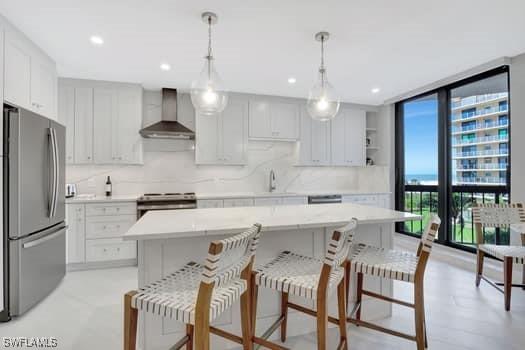 kitchen featuring a kitchen island, hanging light fixtures, stainless steel appliances, wall chimney range hood, and white cabinetry