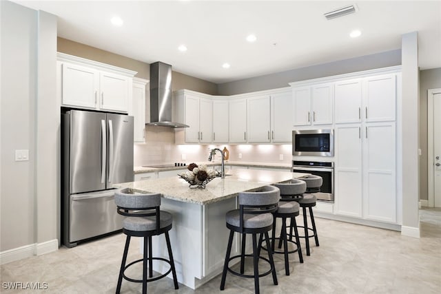 kitchen featuring a kitchen island with sink, wall chimney exhaust hood, a kitchen bar, white cabinetry, and stainless steel appliances