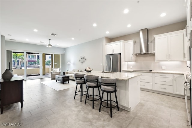 kitchen featuring stainless steel refrigerator, wall chimney range hood, a kitchen breakfast bar, light stone counters, and a center island with sink