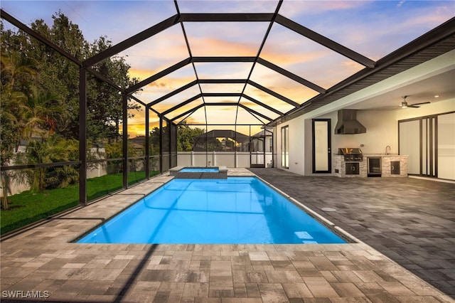 pool at dusk featuring glass enclosure, exterior kitchen, a grill, a patio area, and an in ground hot tub