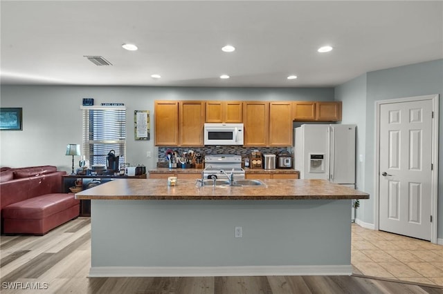 kitchen with sink, backsplash, an island with sink, light hardwood / wood-style floors, and white appliances