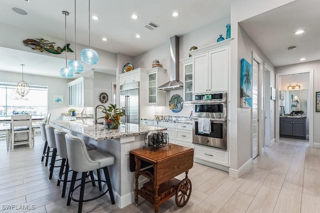 kitchen with appliances with stainless steel finishes, pendant lighting, an island with sink, a kitchen breakfast bar, and wall chimney exhaust hood