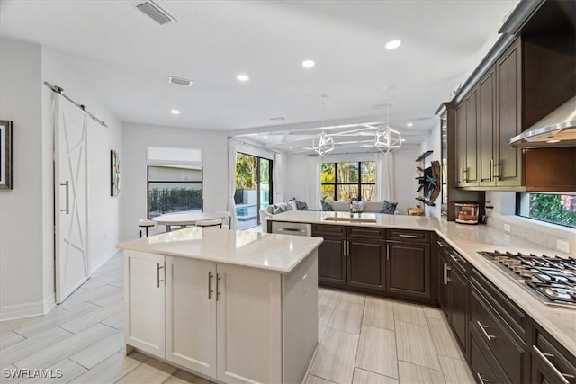 kitchen featuring appliances with stainless steel finishes, wall chimney exhaust hood, dark brown cabinetry, a kitchen island, and hanging light fixtures