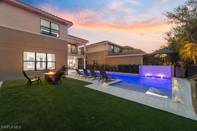 pool at dusk with pool water feature, a patio area, and a lawn