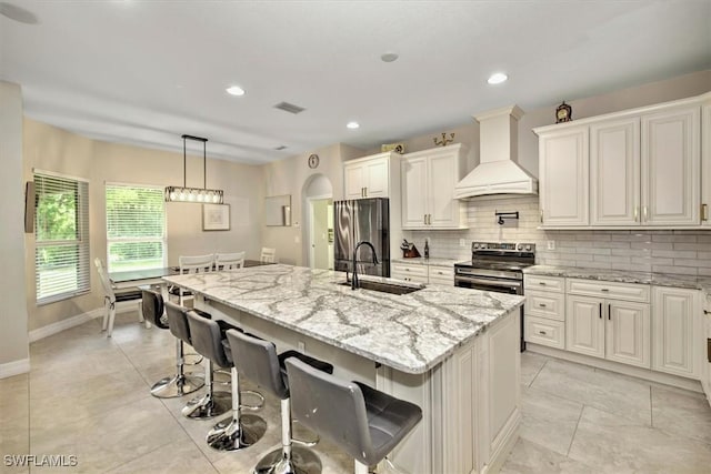 kitchen with appliances with stainless steel finishes, an island with sink, sink, light stone counters, and custom range hood