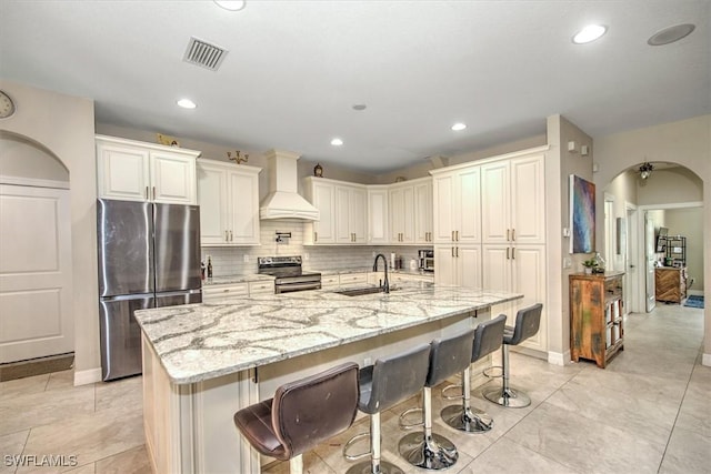 kitchen with appliances with stainless steel finishes, light stone countertops, custom range hood, and a center island with sink