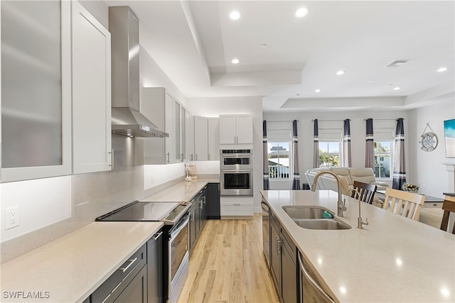 kitchen featuring wall chimney range hood, sink, light stone countertops, appliances with stainless steel finishes, and a tray ceiling