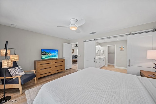 bedroom with light wood-style flooring, a barn door, a ceiling fan, connected bathroom, and baseboards