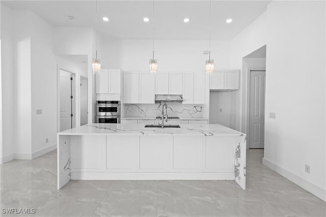 kitchen featuring light stone counters, stainless steel double oven, a large island with sink, decorative light fixtures, and white cabinetry