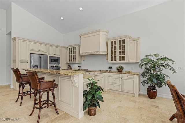 kitchen featuring light stone counters, cream cabinets, a kitchen bar, custom range hood, and appliances with stainless steel finishes