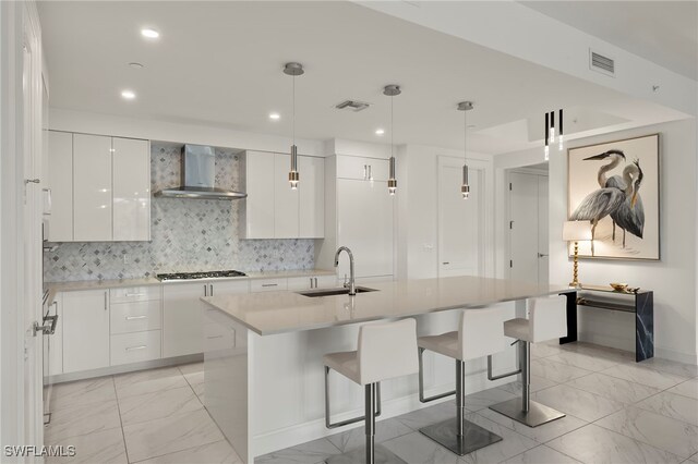 kitchen featuring decorative light fixtures, sink, white cabinets, wall chimney range hood, and a center island with sink