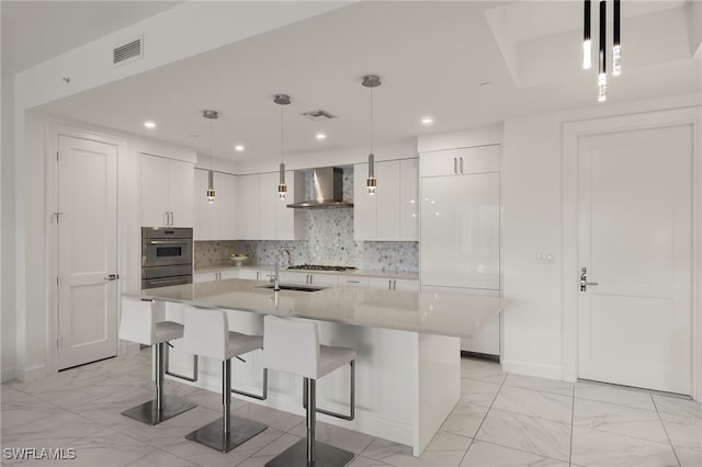 kitchen with hanging light fixtures, a kitchen island with sink, wall chimney range hood, and white cabinets