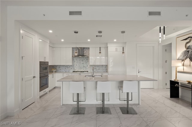 kitchen with white cabinetry, pendant lighting, an island with sink, and wall chimney range hood