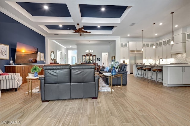 living room with coffered ceiling, ceiling fan with notable chandelier, sink, crown molding, and beam ceiling