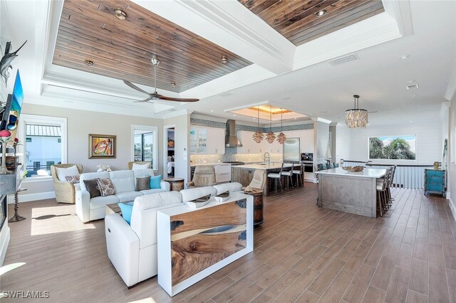 living room with a raised ceiling, wood-type flooring, wood ceiling, ceiling fan with notable chandelier, and ornamental molding