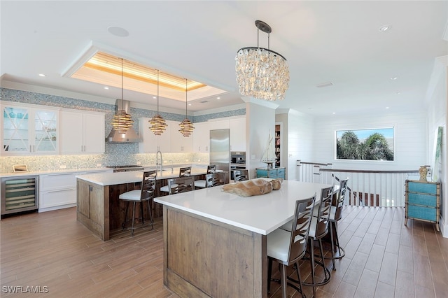 kitchen with white cabinets, a tray ceiling, a large island, and wall chimney range hood