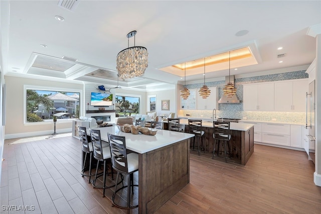 kitchen featuring pendant lighting, a kitchen island with sink, a raised ceiling, wall chimney range hood, and white cabinetry