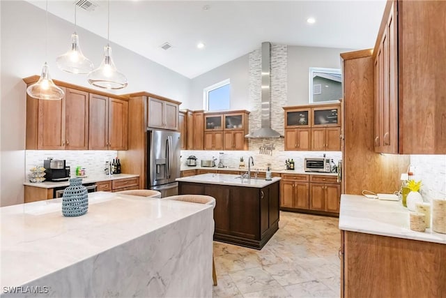 kitchen featuring stainless steel fridge with ice dispenser, a kitchen island with sink, light stone countertops, pendant lighting, and wall chimney range hood