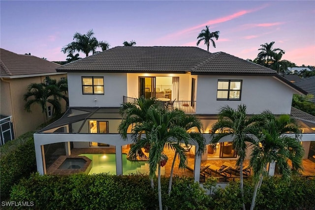 back of property at dusk featuring a balcony, stucco siding, a tile roof, and an in ground hot tub