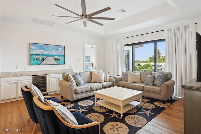 living room featuring sink, ceiling fan, light wood-type flooring, a tray ceiling, and beverage cooler