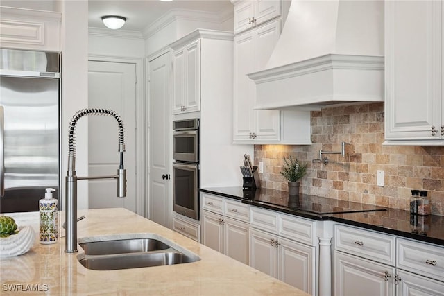 kitchen with appliances with stainless steel finishes, premium range hood, sink, dark stone countertops, and white cabinetry