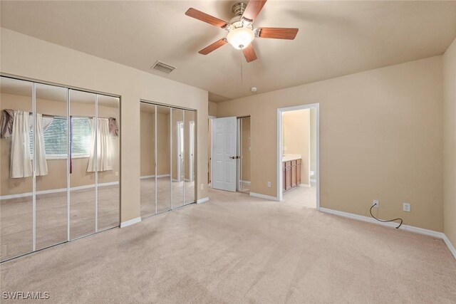 unfurnished bedroom featuring ceiling fan, ensuite bathroom, light colored carpet, and two closets