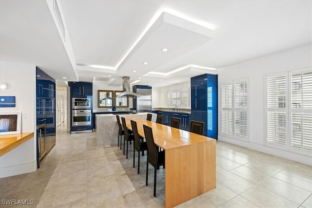 kitchen featuring blue cabinetry, island exhaust hood, a tray ceiling, and a kitchen breakfast bar