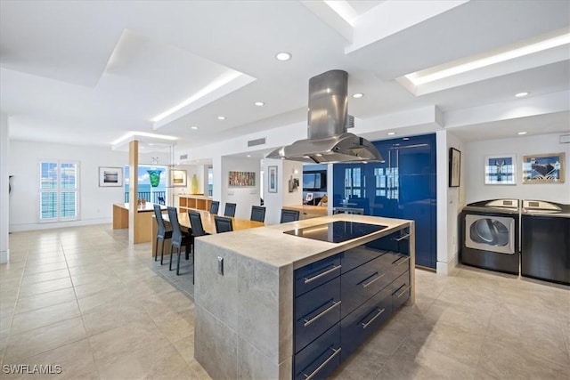 kitchen featuring light tile patterned flooring, washing machine and clothes dryer, a large island with sink, black electric cooktop, and island exhaust hood